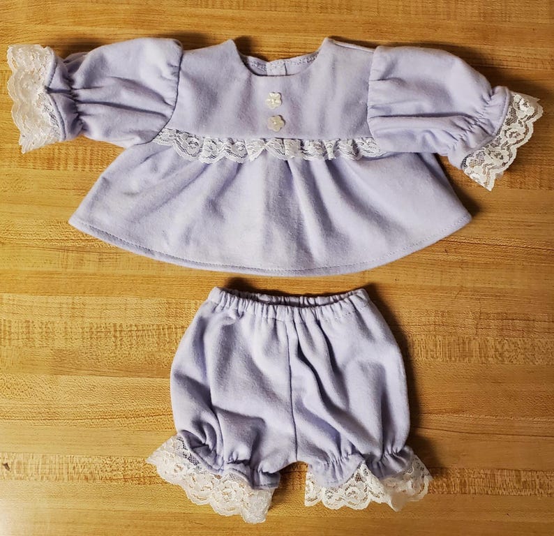 May include: A light purple baby doll outfit, comprising a top with lace trim on the sleeves and bodice, and matching bloomers with lace edging. The top has small flower-shaped buttons.