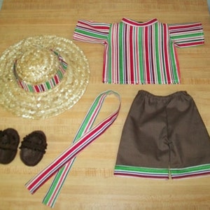 Puede incluir: Un conjunto de ropa para muñeca con un sombrero de paja, una camisa a rayas, pantalones cortos marrones y una cinta roja, verde y blanca. Los pantalones cortos tienen un ribete rojo, verde y blanco.