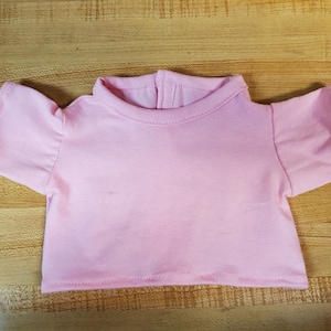 May include: A light pink doll-sized t-shirt with short sleeves and a crew neck. The shirt is made of a soft fabric and has a simple, classic design. The shirt is designed for a doll.