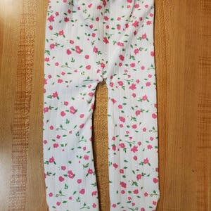 May include: White baby leggings with a pink floral pattern and green leaves. The leggings have a ribbed texture and an elastic waistband. The floral print is evenly distributed across the fabric, creating a delicate and charming design.