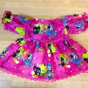 May include: A pink Halloween-themed dress with a lace trim. The dress features a pattern of Mickey Mouse dressed as a witch flying on a broomstick. The dress has a gathered skirt and short sleeves.