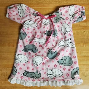 May include: A pink nightdress with a pattern of sleeping cats in grey and white, and pink paw prints. The nightdress has short sleeves, a pink ribbon tie at the neckline, and a white lace trim at the hem.