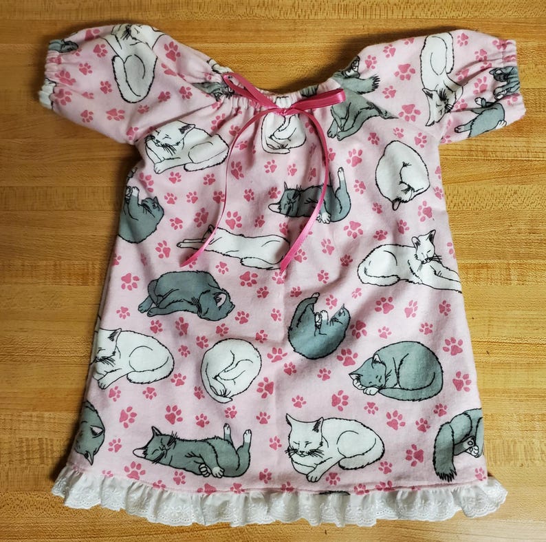 May include: A pink nightdress with a pattern of sleeping cats in grey and white, and pink paw prints. The nightdress has short sleeves, a pink ribbon tie at the neckline, and a white lace trim at the hem.