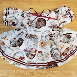 Könnte beinhalten: Ein weißes Kleid mit einem herbstlichen Druckmuster aus Truthähnen, Hähnen, Kürbissen und Erntetexten. Das Kleid hat kurze Puffärmel, einen Rundhalsausschnitt mit einer dunkelroten Schleife und zwei dunkelrote Bandstreifen entlang des Saums.