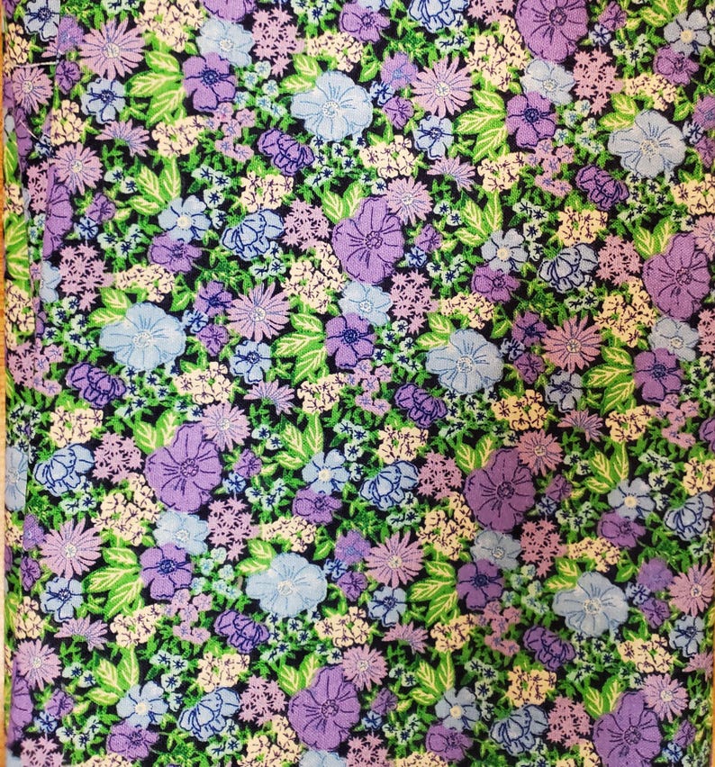May include: A close-up of a fabric with a floral design. The pattern is composed of various flowers in shades of purple, blue, and white, set against a dark green backdrop. Green leaves are scattered throughout, creating a detailed and colourful textile.