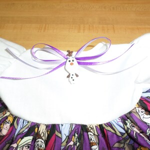 May include: A white dress with a purple and character-themed skirt. The dress features a white bodice with puff sleeves and a purple ribbon bow with an Olaf button. The skirt has a pattern of cartoon characters.