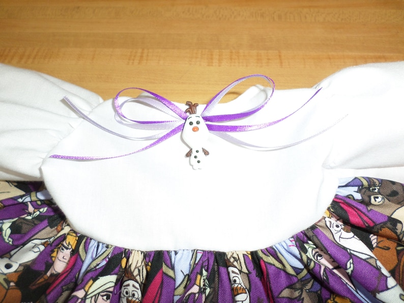 May include: A white dress with a purple and character-themed skirt. The dress features a white bodice with puff sleeves and a purple ribbon bow with an Olaf button. The skirt has a pattern of cartoon characters.