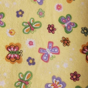 May include: A close-up of a yellow fabric featuring a repeating pattern of butterflies and flowers. The butterflies are in shades of orange, purple, pink, and green. The flowers are pink, orange, and purple. The background is a solid yellow.