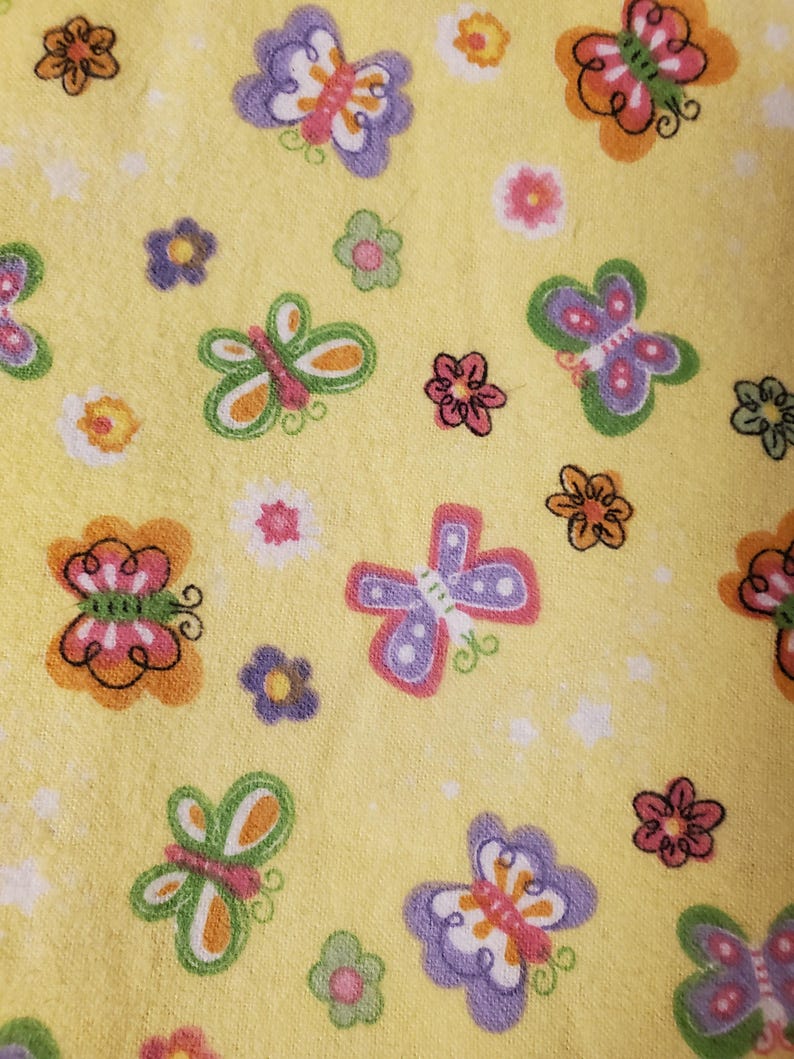 May include: A close-up of a yellow fabric featuring a repeating pattern of butterflies and flowers. The butterflies are in shades of orange, purple, pink, and green. The flowers are pink, orange, and purple. The background is a solid yellow.