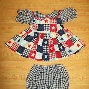 May include: A patriotic baby dress and bloomers set. The dress features a patchwork design with red, white, and blue squares, accented with star patterns. The sleeves and collar are a blue and white gingham. The bloomers match the gingham pattern.