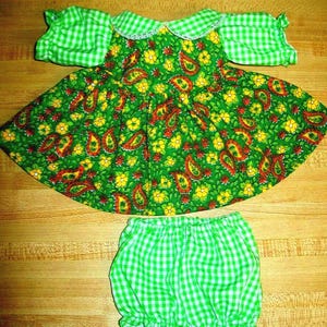 May include: A doll dress and bloomers set. The dress has a green paisley floral pattern with yellow flowers and red accents. The sleeves and collar are green and white gingham. The matching bloomers are also green and white gingham.