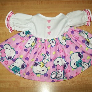 May include: A pink and white dress with a Snoopy print. The dress has a white bodice with pink buttons and pink trim on the sleeves. The skirt is pink with a Snoopy and heart print.