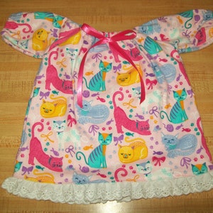May include: A pink and white dress with a cat print and a pink ribbon. The dress has a white lace trim and short sleeves.