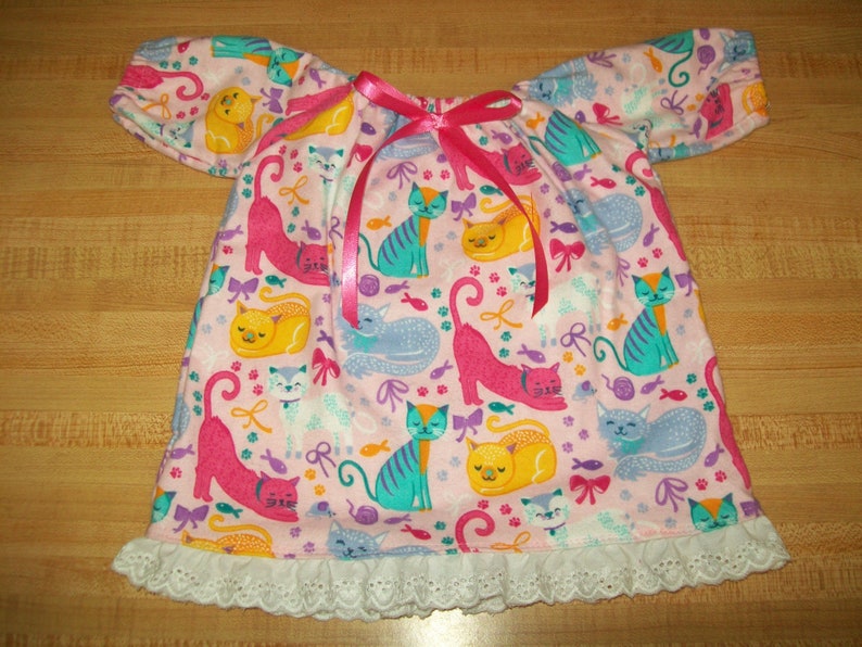 May include: A pink cat-patterned dress with short sleeves and a white lace trim. The dress features a repeating pattern of colourful cats, fish, and bows. A pink ribbon is tied at the neckline.