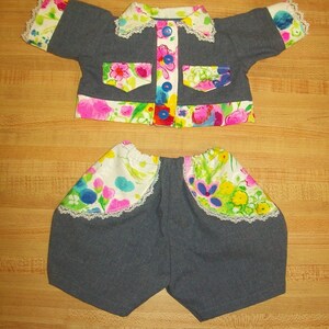 Op de afbeelding: Een denim jack en shorts set voor een pop. Het jack heeft een witte kraag met kant en bloemenstof aan de voor- en achterkant. De shorts hebben een bloemenstof rand en kant rond de bovenkant.