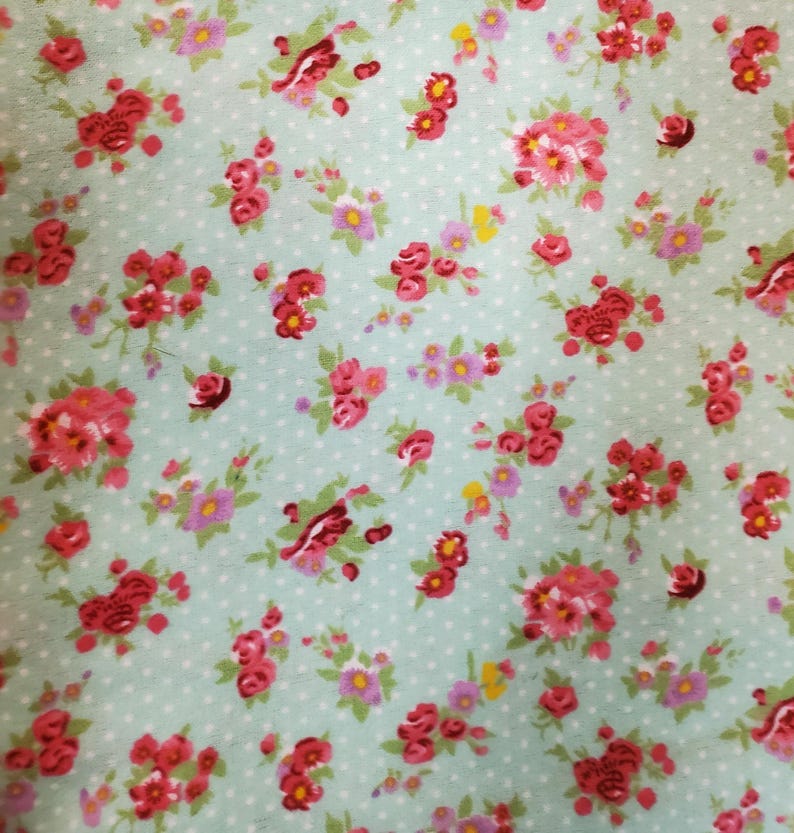 May include: A floral fabric with a repeating pattern of red and pink roses, small purple flowers, and green leaves. The design is set against a light blue backdrop with small white polka dots.