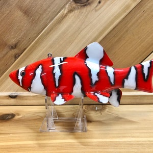 May include: A red, white, and black painted fish lure with a realistic design. The lure is on a clear plastic stand.