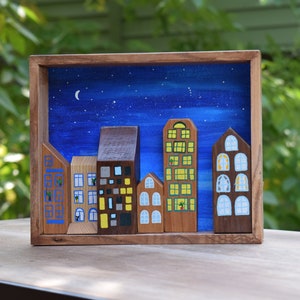 May include: A wooden shadow box frame containing five distinct wooden building cutouts arranged to form a city skyline. The background is painted with a dark blue night sky featuring a crescent moon and stars. Each building has painted windows.