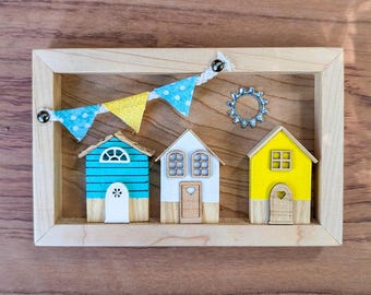 May include: A wooden frame with three miniature houses inside. The houses are painted in blue, white, and yellow with white trim. A string of blue and yellow bunting hangs above the houses.