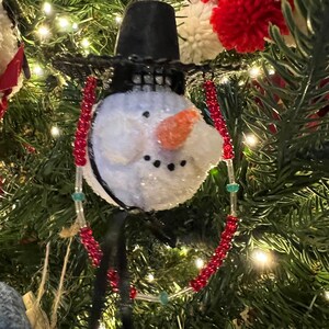 K&ouml;nnte beinhalten: Ein Schneemann-Weihnachtsbaumschmuck mit einem schwarzen Hut und einer roten und wei&szlig;en Perlenkette. Der Schneemann hat einen wei&szlig;en, flauschigen K&ouml;rper, einen schwarzen Mund und eine orangefarbene Karottennase.