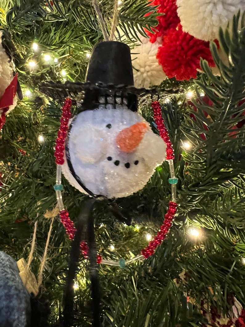 K&ouml;nnte beinhalten: Ein Schneemann-Weihnachtsbaumschmuck mit einem schwarzen Hut und einer roten und wei&szlig;en Perlenkette. Der Schneemann hat einen wei&szlig;en, flauschigen K&ouml;rper, einen schwarzen Mund und eine orangefarbene Karottennase.