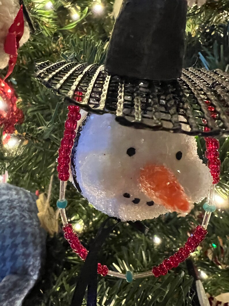 K&ouml;nnte beinhalten: Ein wei&szlig;er Schneemann-Anh&auml;nger mit einem schwarzen Hut und roten und t&uuml;rkisfarbenen Perlen. Der Schneemann hat eine Karottennase und schwarze Augen.