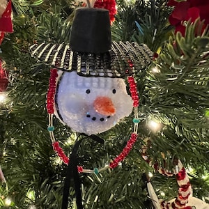 K&ouml;nnte beinhalten: Ein wei&szlig;er Schneemann-Anh&auml;nger mit einem schwarzen Hut und einem schwarz-wei&szlig; gestreiften Rand. Der Schneemann hat rote und t&uuml;rkisfarbene Perlen um den Hals.
