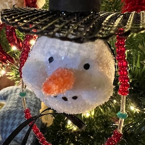 K&ouml;nnte beinhalten: Ein Schneemann-Anh&auml;nger mit einem schwarzen Hut und einer roten Perlenkette. Der Schneemann hat ein wei&szlig;es, flauschiges Gesicht mit einer Karottennase und schwarzen Augen.