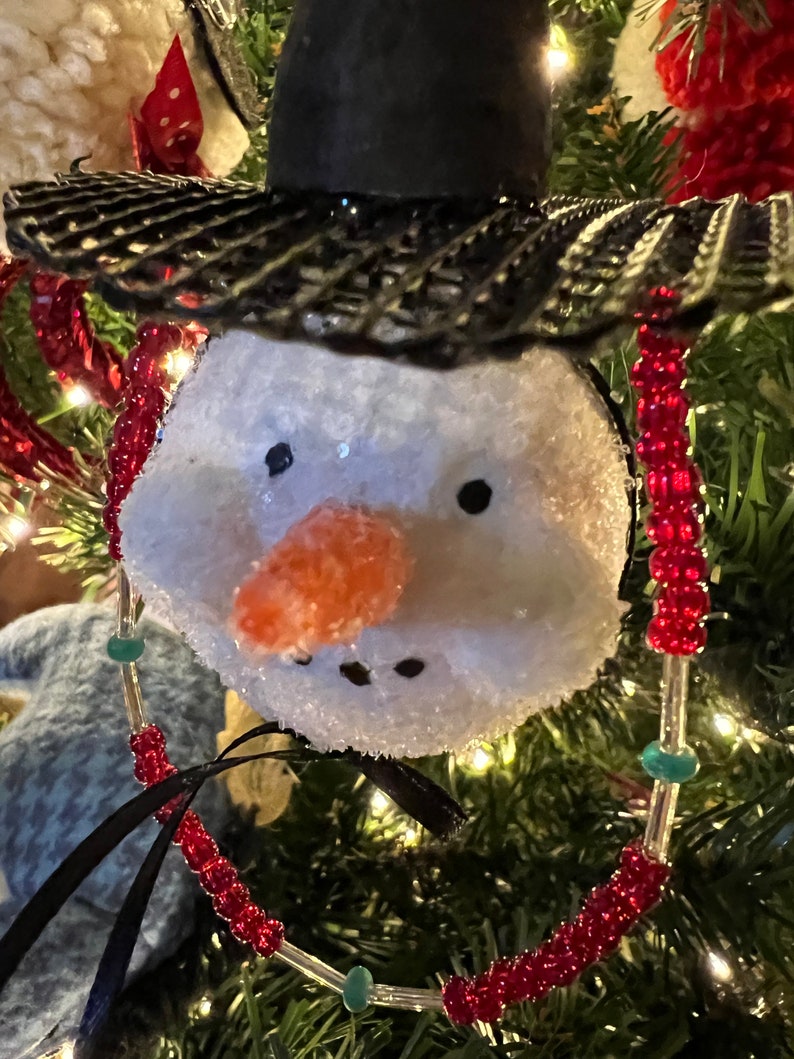 K&ouml;nnte beinhalten: Ein Schneemann-Anh&auml;nger mit einem schwarzen Hut und einer roten Perlenkette. Der Schneemann hat ein wei&szlig;es, flauschiges Gesicht mit einer Karottennase und schwarzen Augen.
