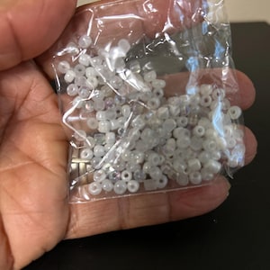 May include: A clear plastic bag filled with small, round, white beads. The beads appear to be made of glass or a similar material, with a slightly iridescent sheen. The beads are held in a person's hand.
