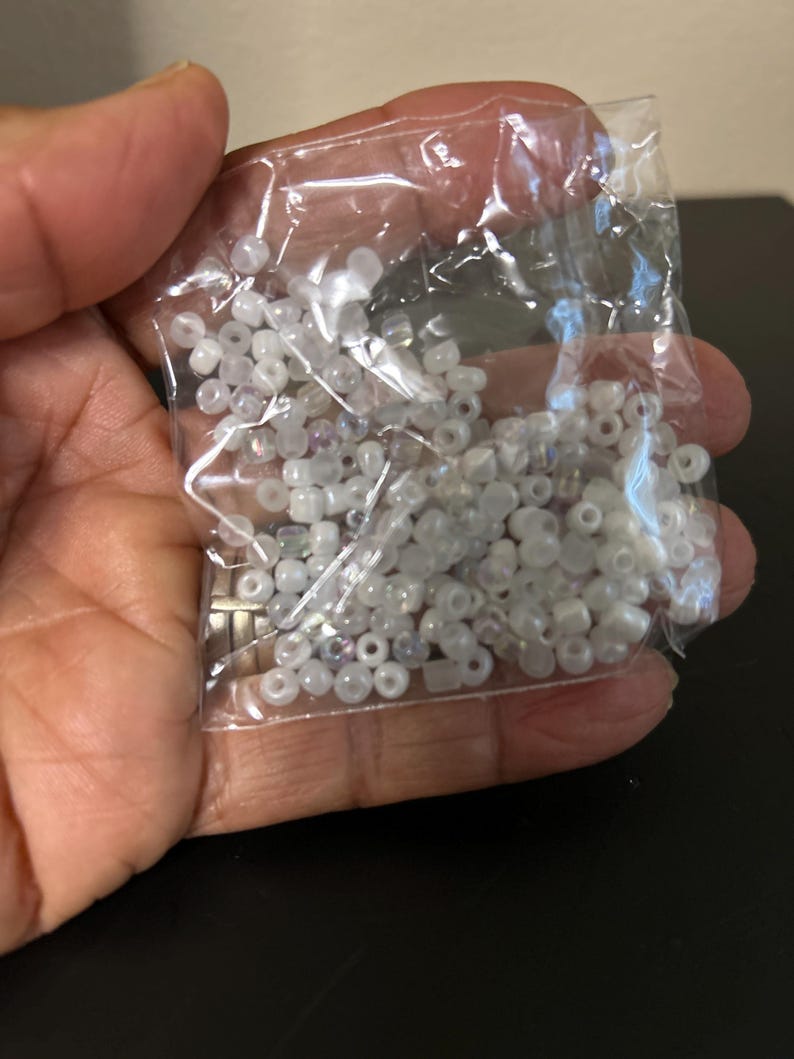 May include: A clear plastic bag filled with small, round, white beads. The beads appear to be made of glass or a similar material, with a slightly iridescent sheen. The beads are held in a person's hand.