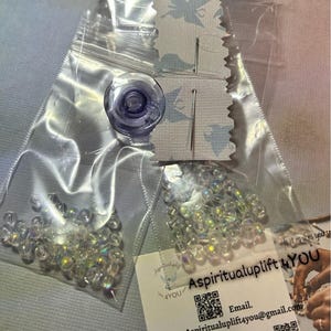 May include: Clear plastic bags containing craft supplies. One bag holds iridescent beads, another contains a purple bobbin, needles, and thread. A small tag with the text "Aspiritualuplift 4YOU" and contact information is also visible.