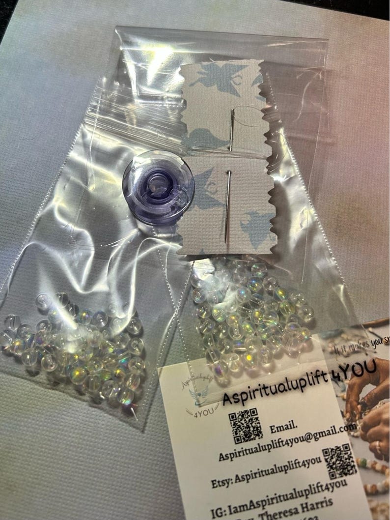May include: Clear plastic bags containing craft supplies. One bag holds iridescent beads, another contains a purple bobbin, needles, and thread. A small tag with the text "Aspiritualuplift 4YOU" and contact information is also visible.