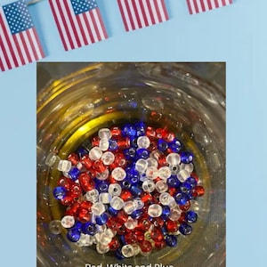 May include: A glass jar filled with red, white, and blue beads. Above the jar, small American flags are strung together. The text "Red, White and Blue" is at the bottom of the jar. The image has a patriotic theme.