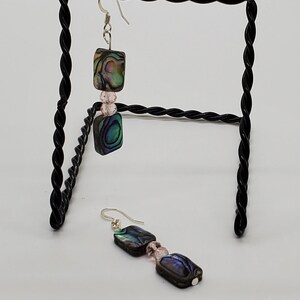May include: A pair of earrings with iridescent abalone shell squares and pink beads hanging from silver hooks.
