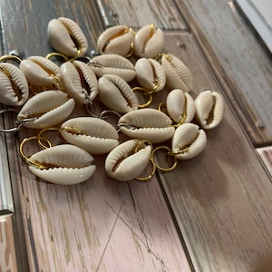 May include: A collection of off-white cowrie shells, each individually attached to a small, circular gold-toned metal jump ring. The shells are arranged in a cluster, showcasing their natural ribbed texture and central openings against a surface.