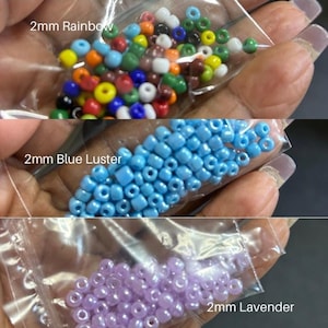May include: Three small plastic bags filled with 2mm beads in rainbow, blue luster, and lavender colors. The rainbow beads are a mix of colors, the blue beads are a light blue, and the lavender beads are a soft purple. The text "2mm Rainbow", "2mm Blue Luster", and "2mm Lavender" are printed on the bags.