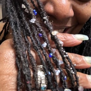 May include: Black dreadlocks adorned with clear, blue, and silver beads and charms. A silver ring with a blue stone is visible. The dreadlocks are styled with various decorative elements.