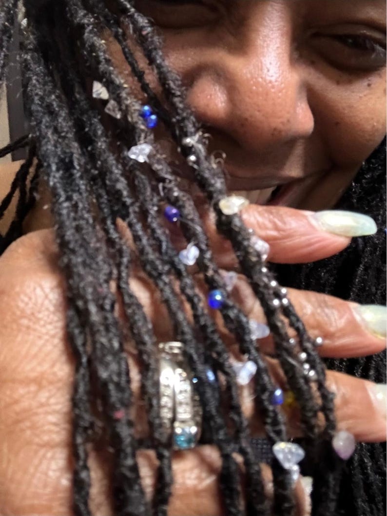 May include: Close-up of dark, textured dreadlocks adorned with clear, blue, and silver beads. A silver ring with a blue stone is visible. The image captures a close-up of the hair and jewelry.