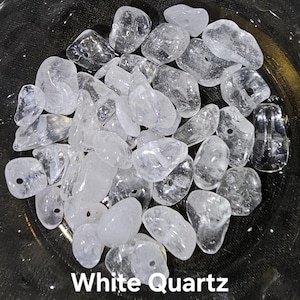 May include: A collection of clear, white quartz stones, some with drilled holes, are displayed in a dark bowl. The stones vary in size and shape, with a translucent appearance. The text "White Quartz" is visible at the bottom of the image.