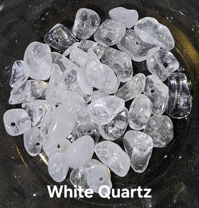 May include: A collection of clear, white quartz stones, some with drilled holes, are displayed in a dark bowl. The stones vary in size and shape, with a translucent appearance. The text "White Quartz" is visible at the bottom of the image.