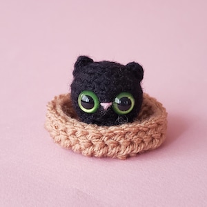 May include: A small, round, black crocheted cat head with large, bright green plastic eyes and a tiny pink stitched nose, resting inside a circular, tan-coloured crocheted base with a textured, braided-style edge.