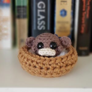 May include: A small brown crocheted monkey head with large black plastic eyes and a cream-colored muzzle, nestled inside a circular tan crocheted basket. The monkey features rounded ears and a textured yarn surface, creating a compact figure.