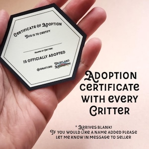 May include: A hexagonal certificate of adoption with black borders and white text. The text reads "Certificate of Adoption" and "This is to certify". The certificate is blank and has a space for the name of the critter and a signature line. The text "Signature: Fairyland Amigurumi" is printed below the signature line. The certificate is held in a hand. The text "Adoption Certificate with Every Critter" is printed below the certificate. The text "* Arrives Blank! If you would like a name added please let me know in message to seller" is printed below the text "Adoption Certificate with Every Critter".