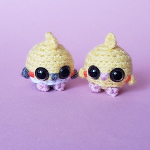 May include: Two handmade, yellow crochet bird-like creatures with large, black eyes and small, pink feet. The birds have small, orange accents and a tuft of yellow yarn on top of their heads. They are set against a soft purple background.