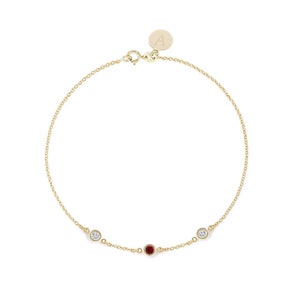May include: A gold chain bracelet with a small red gemstone and two smaller white gemstones. The bracelet has a clasp with a small charm that says "A".
