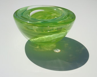 May include: A lime green glass bowl with a thick, rounded rim and a smooth, translucent finish. The side of the bowl features the black text Kosta Boda printed clearly on the surface. The bowl has a circular opening at the top.