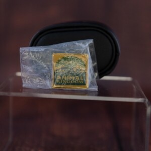 May include: A gold-colored pin with the words "ANIMAL KINGDOM" and a tree design, encased in a clear plastic bag. A black, oval-shaped object is behind the pin. The pin is on a clear, rectangular surface.