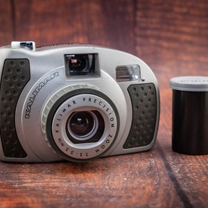May include: A silver and black Kalimar Precision Zoom 35mm film camera with a lens and flash. A black film canister with a gray lid sits to the right of the camera. The camera has a black strap and is on a wooden surface.