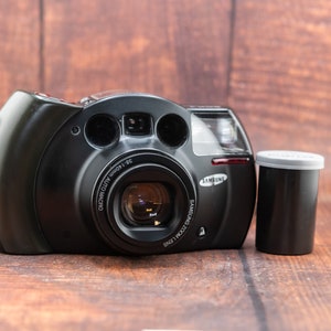 May include: A black Samsung compact film camera with a silver lens and a black film canister.